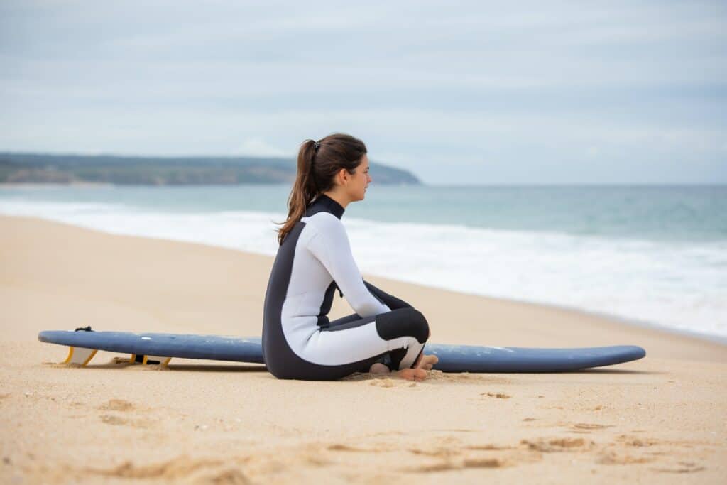 a woman sitting beside a surfboard wearing black and white wetsuit