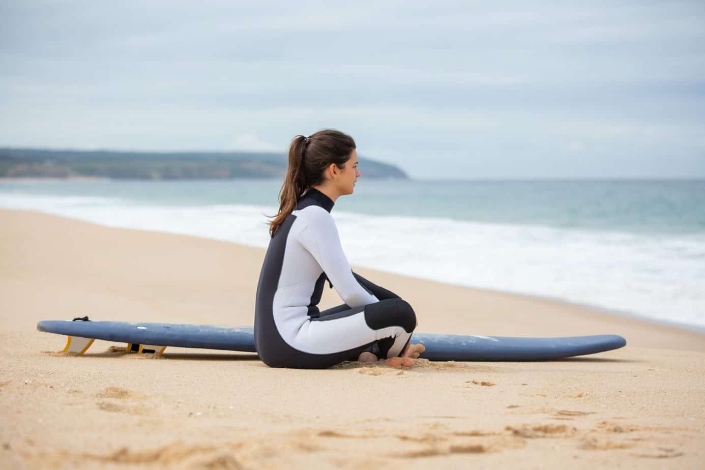 a woman sitting beside a surfboard wearing black and white wetsuit