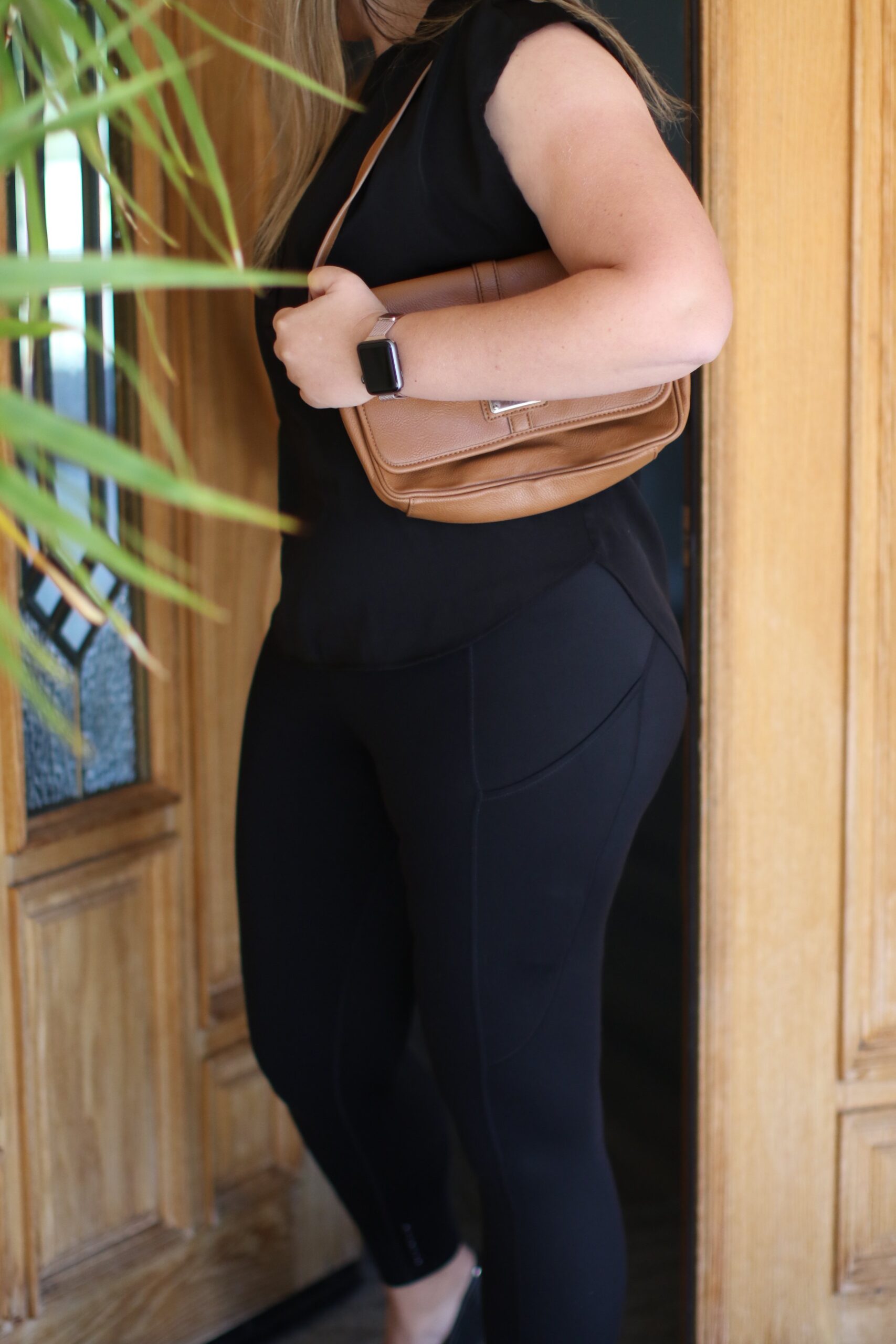 Woman wearing a black blouse and black leggings going outside and closing a brown door 