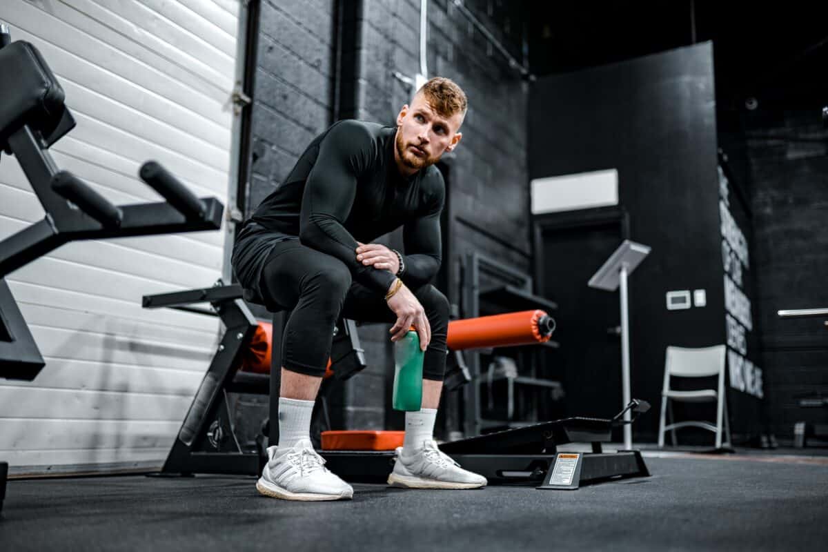 A man with brown hair wears a black rashguard, black shorts, and black compression pants while sitting on a piece of equipment at the gym