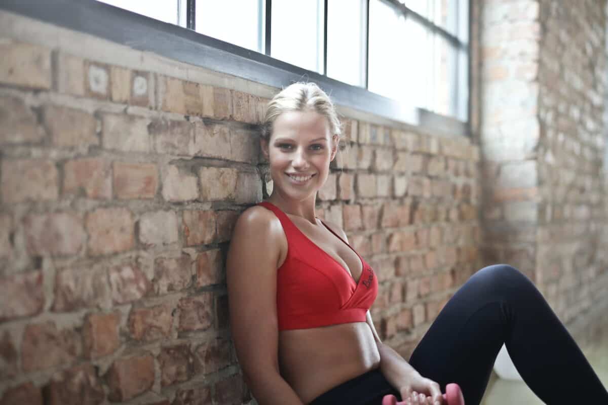 A blonde girl wearing a red sports bra and black leggings sits on the floor while leaning on the bricked wall