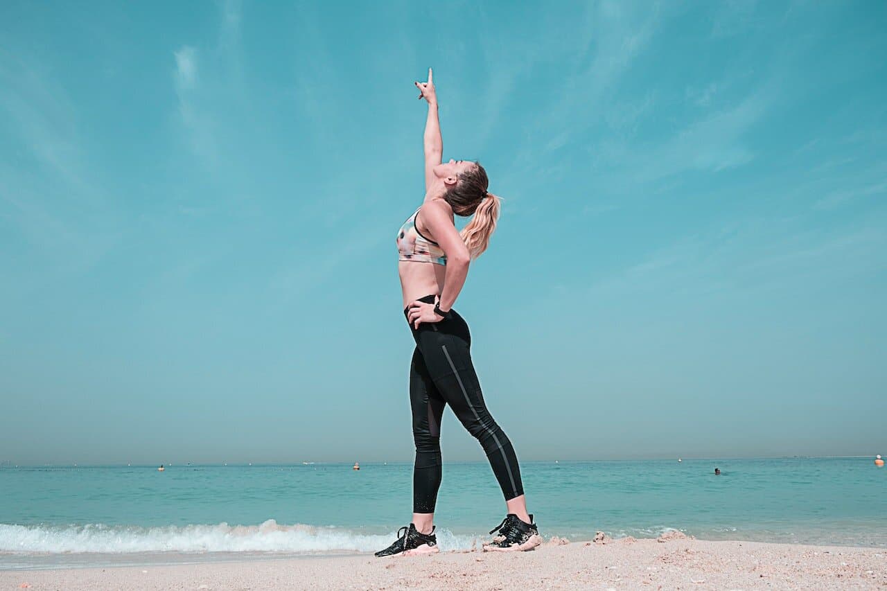 A woman wearing a printed sports bra and black leggings is pointing at the sky using her right hand while standing near the shore