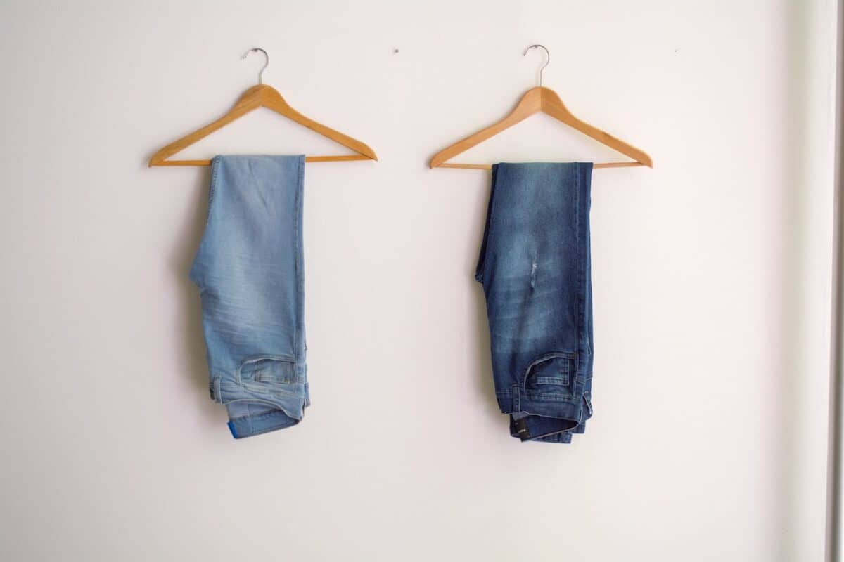 Two denim pants hanging on a wooden hanger placed on a white wall inside a room