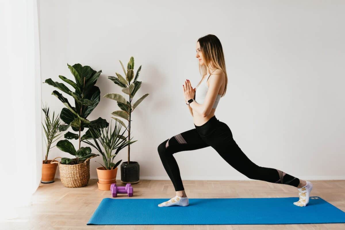 A woman wearing a gray sports bra and black leggings doing yoga poses on a blue yoga mat near purple dumbells and fresh plants in a well-lit room