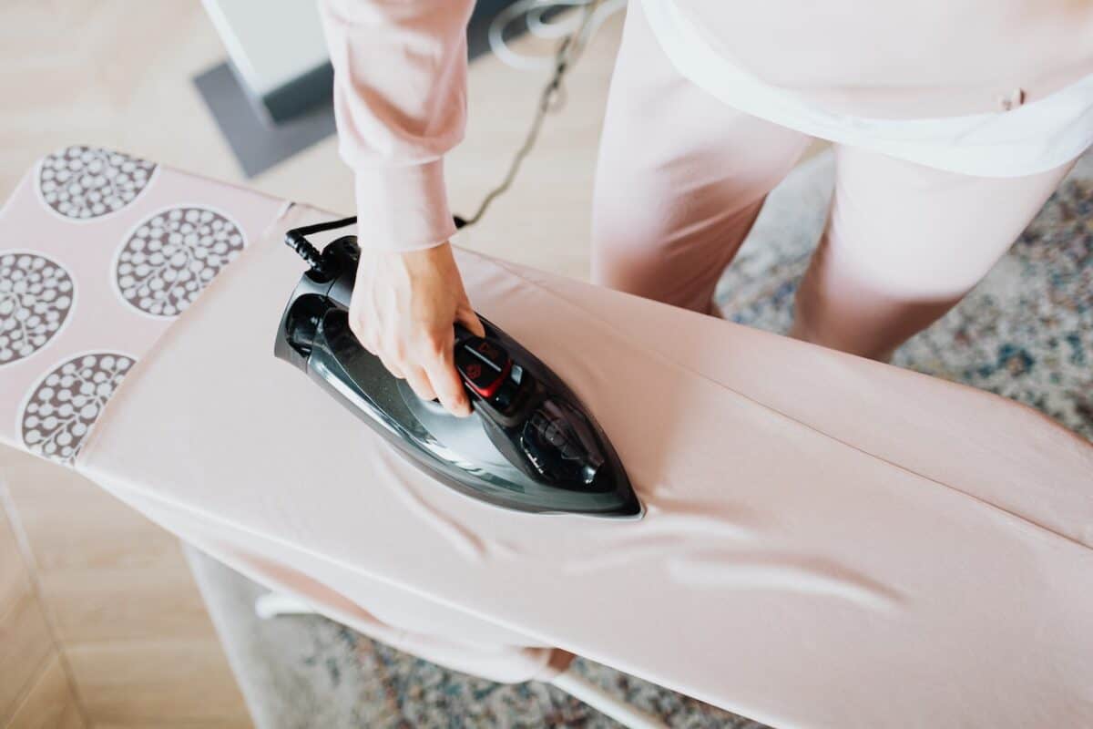 A woman standing on a gray carpet ironing pink clothing using a black iron and printed ironing board in a room