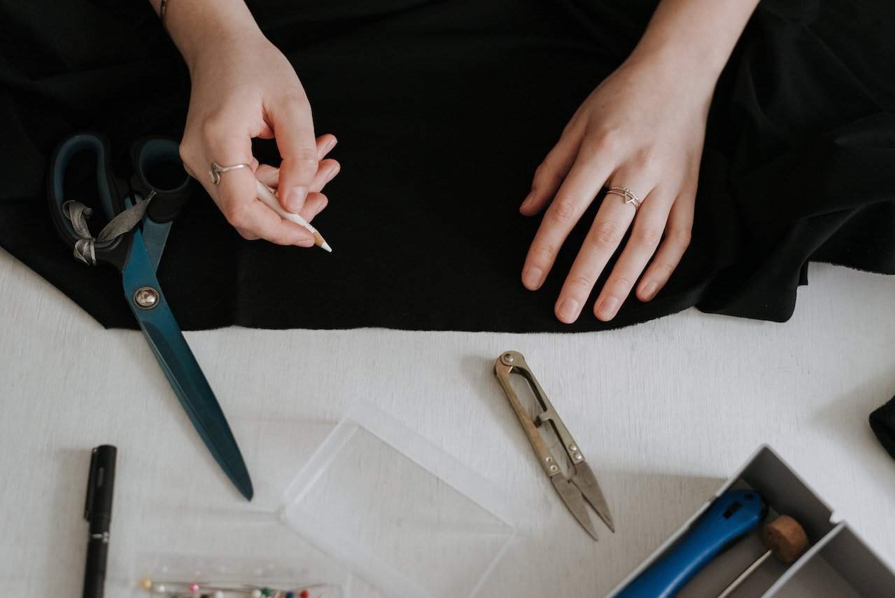 A person wearing two silver rings holds a white pencil to mark a black cloth near two sharp cutting tools on a white wooden table