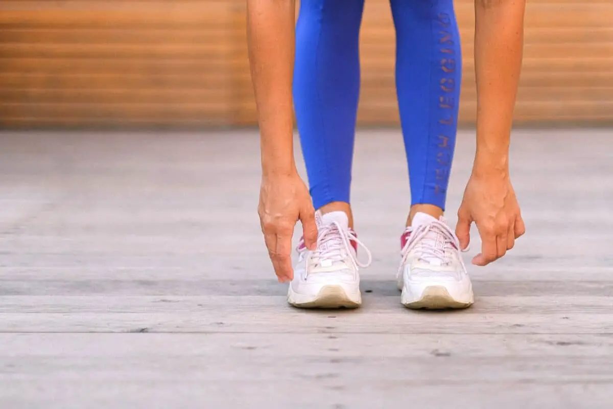 A woman wearing blue leggings stretches her hands down towards her white running shoes while standing on a wooden floor