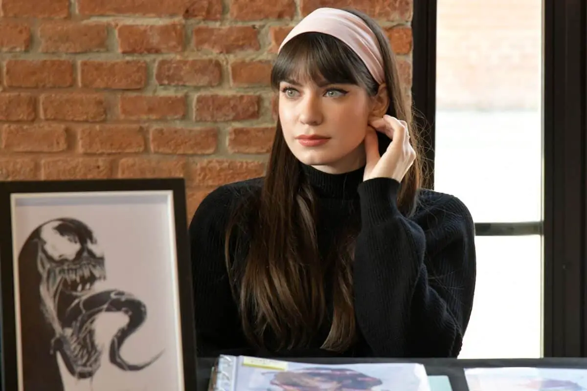 A woman with brown hair wears a pink headband and black turtleneck shirt while sitting near a brick wall