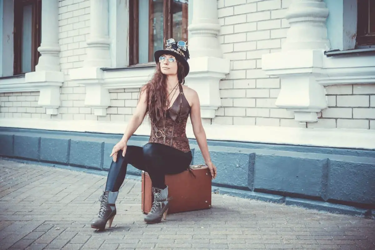 A woman with brown hair wearing a black hat s sitting in a brown suitcase placed on a pavement