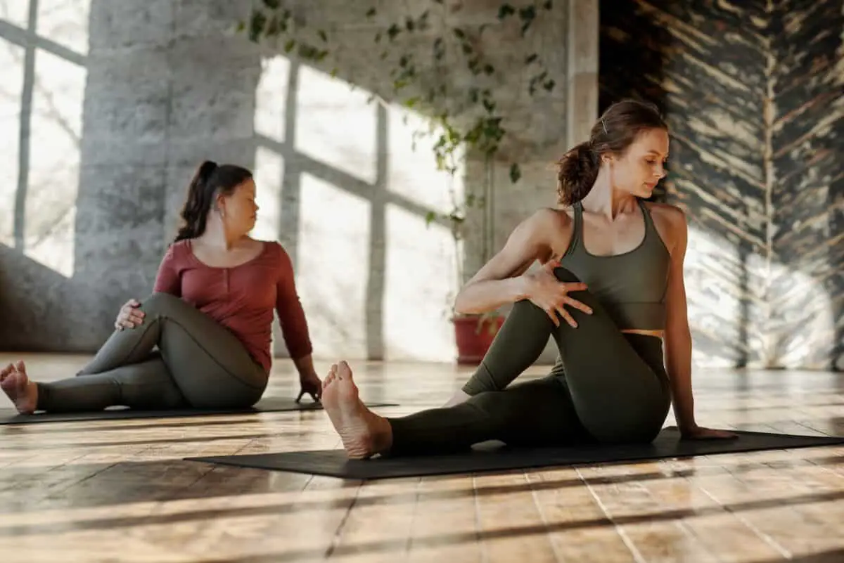 Two women wearing gray leggings doing stretches on a black yoga mat inside a yoga studio