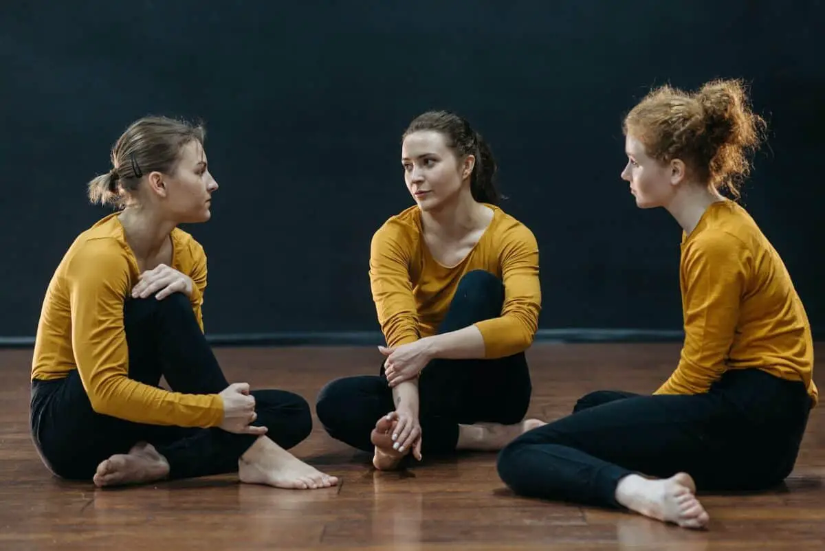 Three women wearing yellow long sleeves tops and black leggings sitting on a brown wooden floor in a dance studio