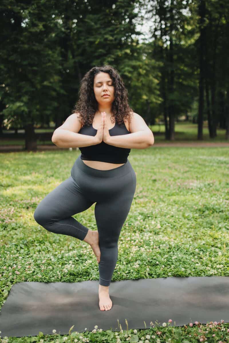 A woman wears a black sports bra and gray leggings doing a yoga pose on a gray yoga mat at the park