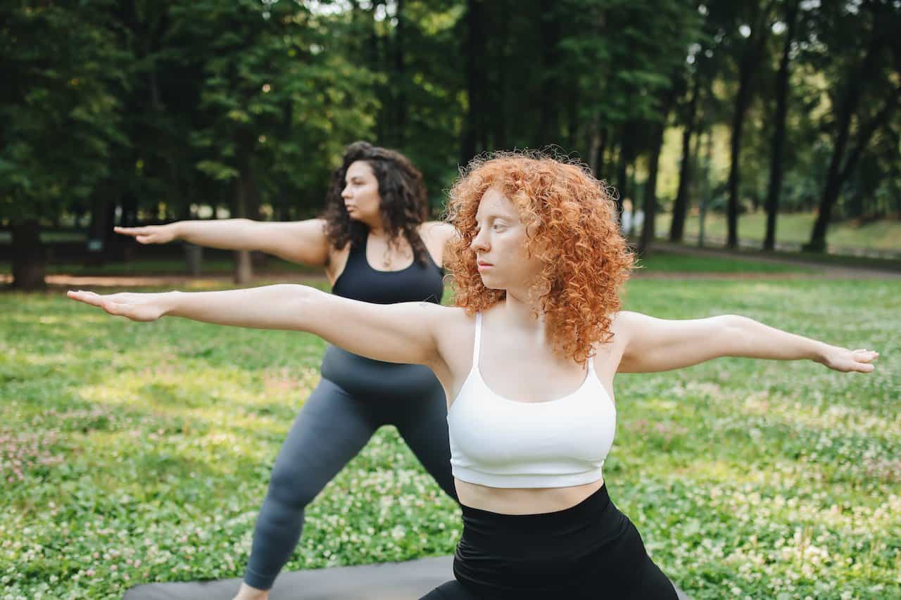 Two women doing a yoga pose wearing dark-colored leggings standing on a gray mat in the park