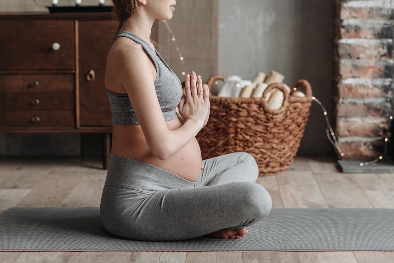 A pregnant woman wearing a gray tank top and gray leggings is seated on a yoga mat doing meditation in her apartment