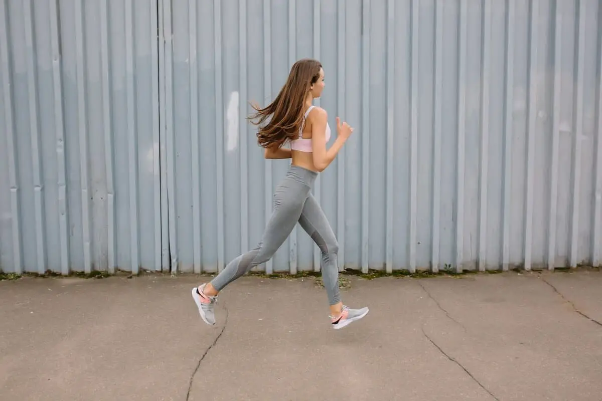 A woman with brown hair wears a pink sports bra and gray leggings while running near a gray wall outdoors