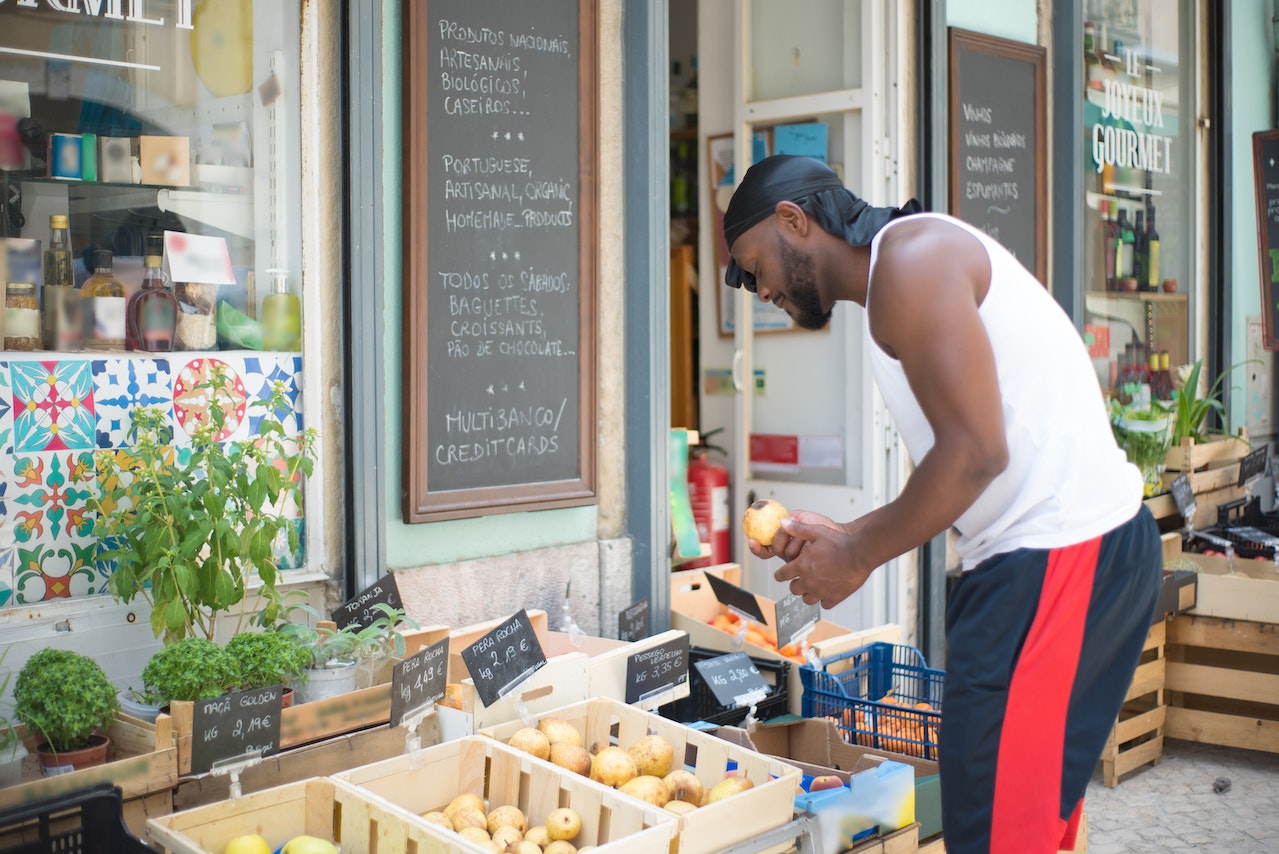 A man in a white sando and black shorts wears a black wave cap while looking a fruits outside the grocery store