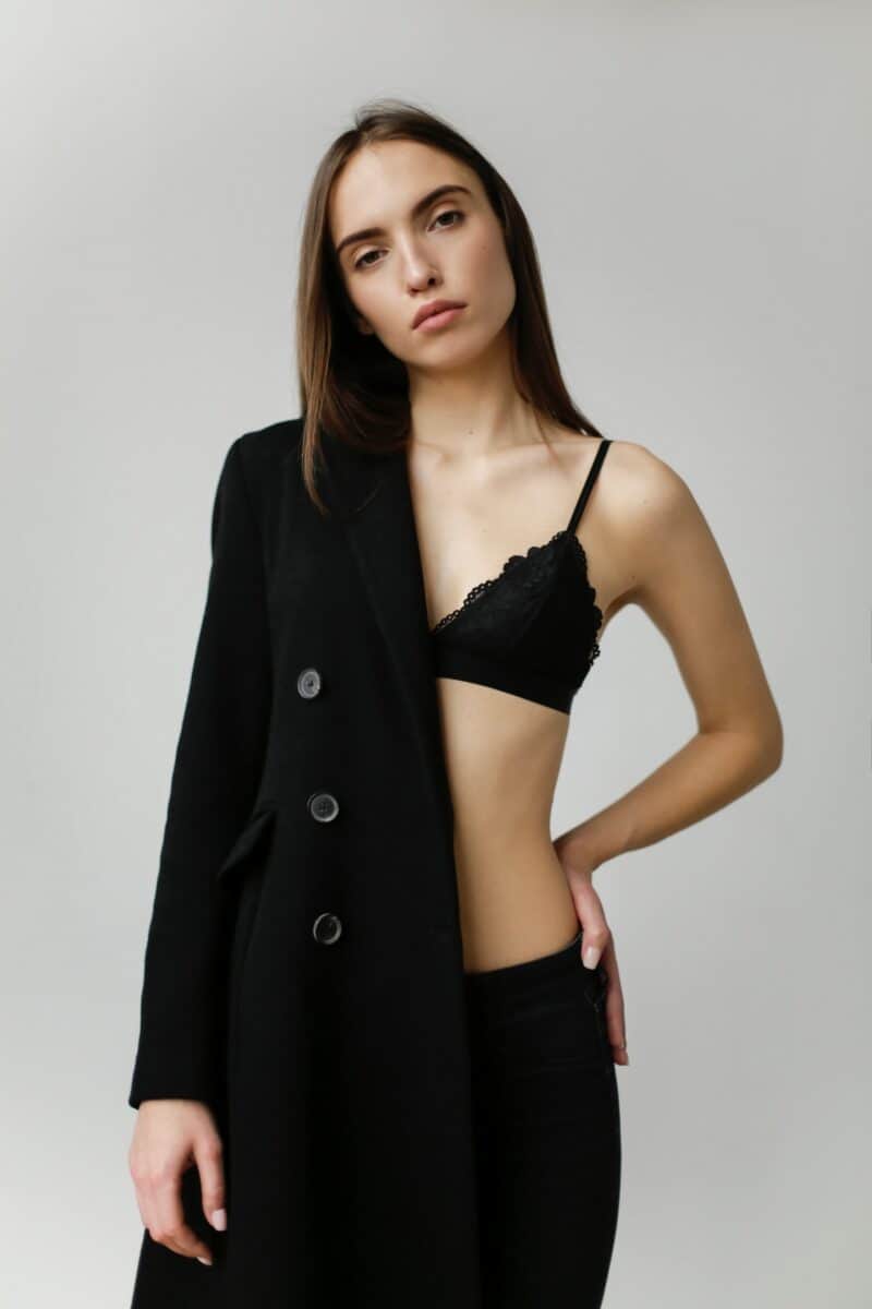 A woman with long brown hair wears a black coat and a black bra standing near a gray wall