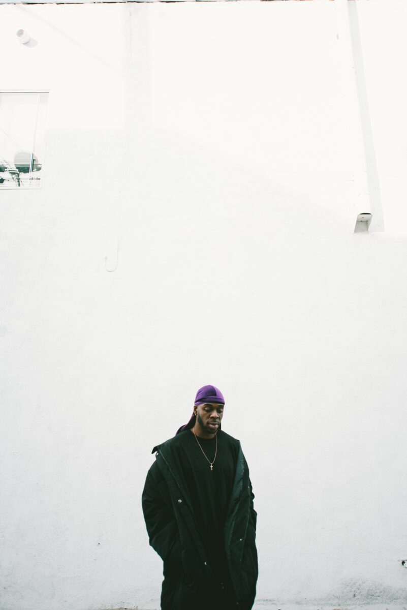 A man in full-black attire wearing a purple wave cap and gold necklace standing near a white wall