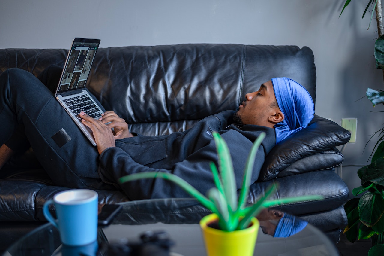 A man in a black jacket and black pants wears a blue wave cap and using a laptop while lying on a black couch