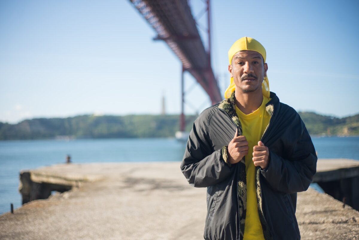 A man wearing a black jacket over a yellow shirt is standing near a body of water overlooking a long bridge