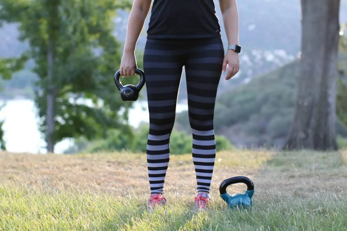 A woman in black and gray striped leggings wearing a smartwatch and holding a black kettlebell in the grass field