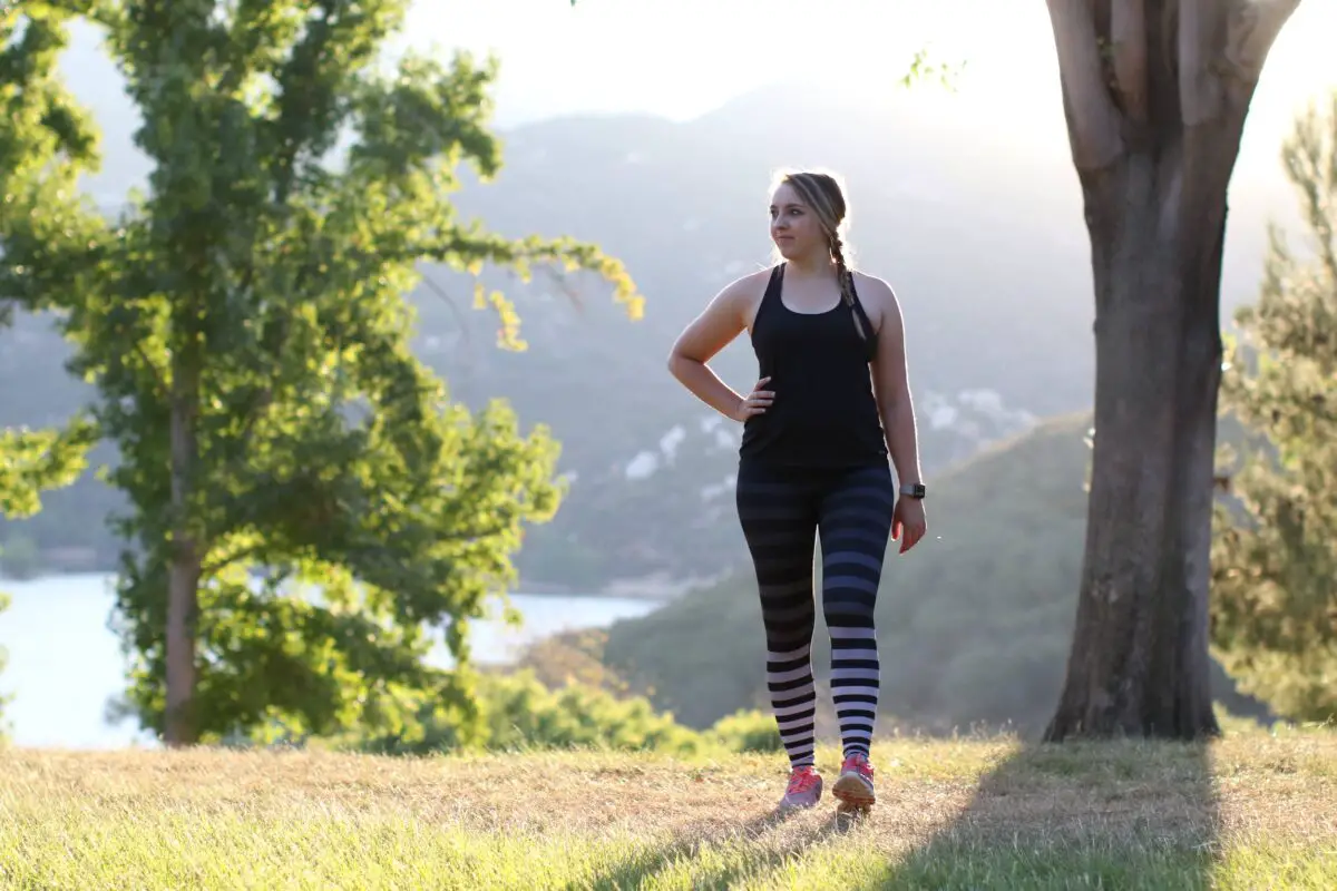 A woman in a black tank top and striped leggings wearing purple rubber shoes walks into the grass field