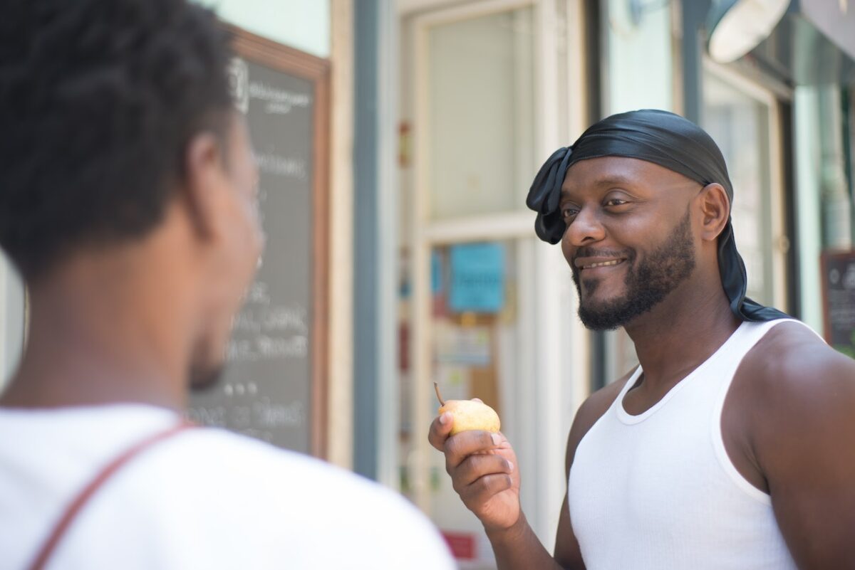 A man wearing a white tank top is showing a fresh pear to a person in a white shirt while standing outside a grocery store