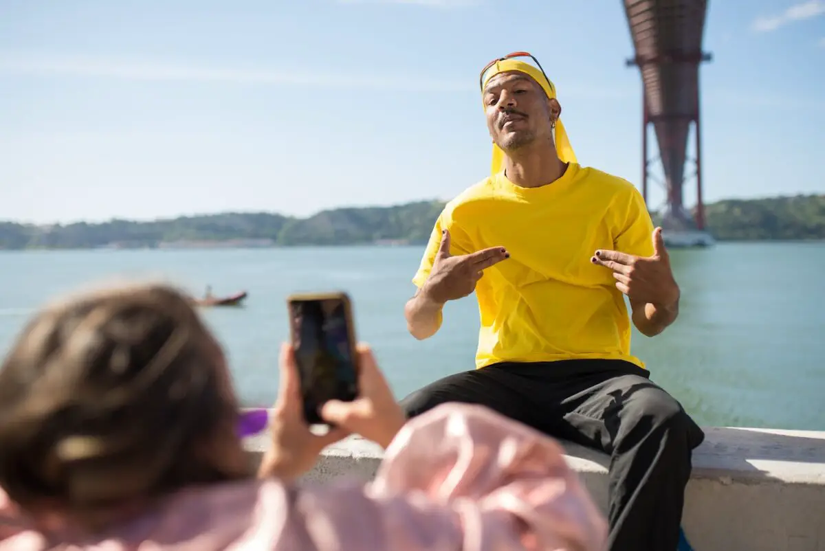 A person wearing a pink jacket is taking a photo of a man in a yellow shirt and yellow durag near a body of water