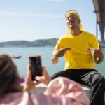A person wearing a pink jacket is taking a photo of a man in a yellow shirt and yellow durag near a body of water