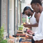 A man in a white tank top and black durag while the other man wears a white long sleeves sweatshirt looking for fruits outside the grocery store