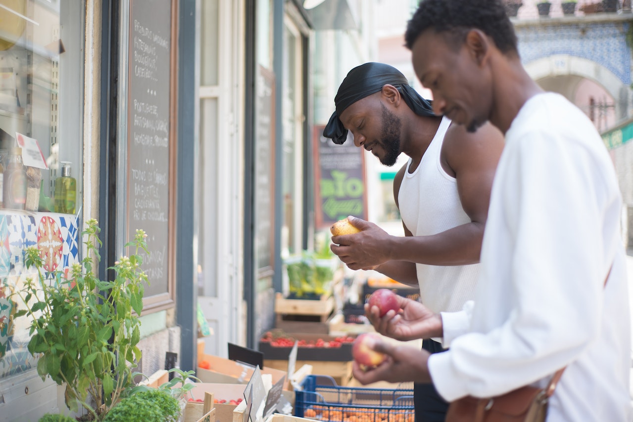 A man in a white tank top and black durag while the other man wears a white long sleeves sweatshirt looking for fruits outside the grocery store