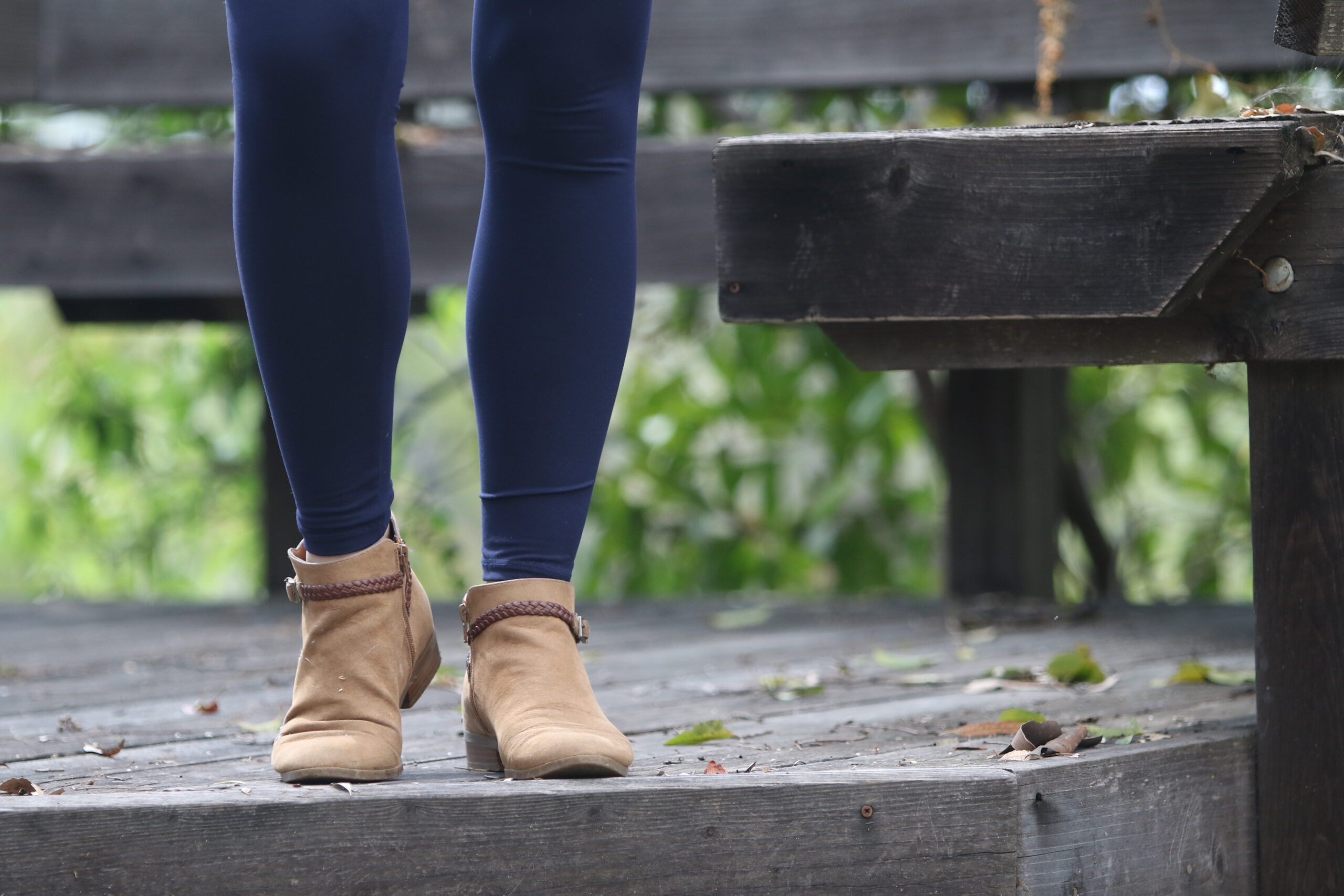 A person in blue leggings and brown ankle boots stands on a black wooden plank in the park