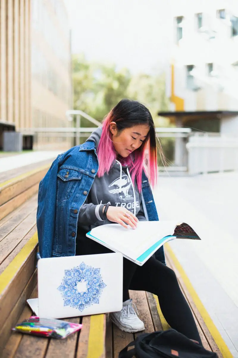 A woman in a gray sweatshirt and blue denim jacket wearing black leggings and white rubber shoes holding an art workbook outside the school