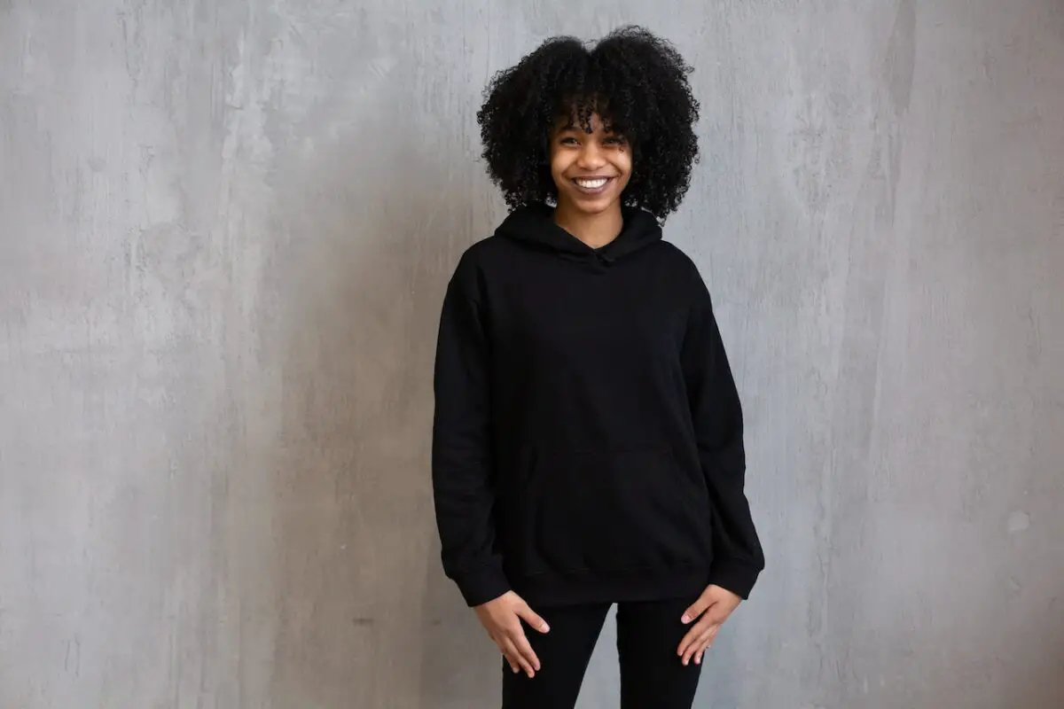 A woman smiling widely, wearing a black sweatshirt and black leggings, standing near a gray wall
