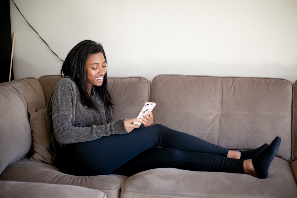 Woman in gray velvet long sleeves and black leggings wearing black socks holding a smartphone while sitting on a brown couch