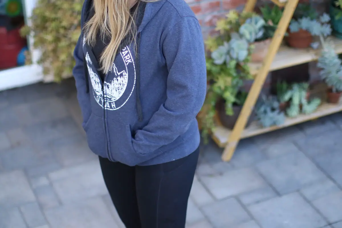 Woman wearing a blue hoodie with her hands in the sweatshirt pockets standing outside on gray pavers in the backyard