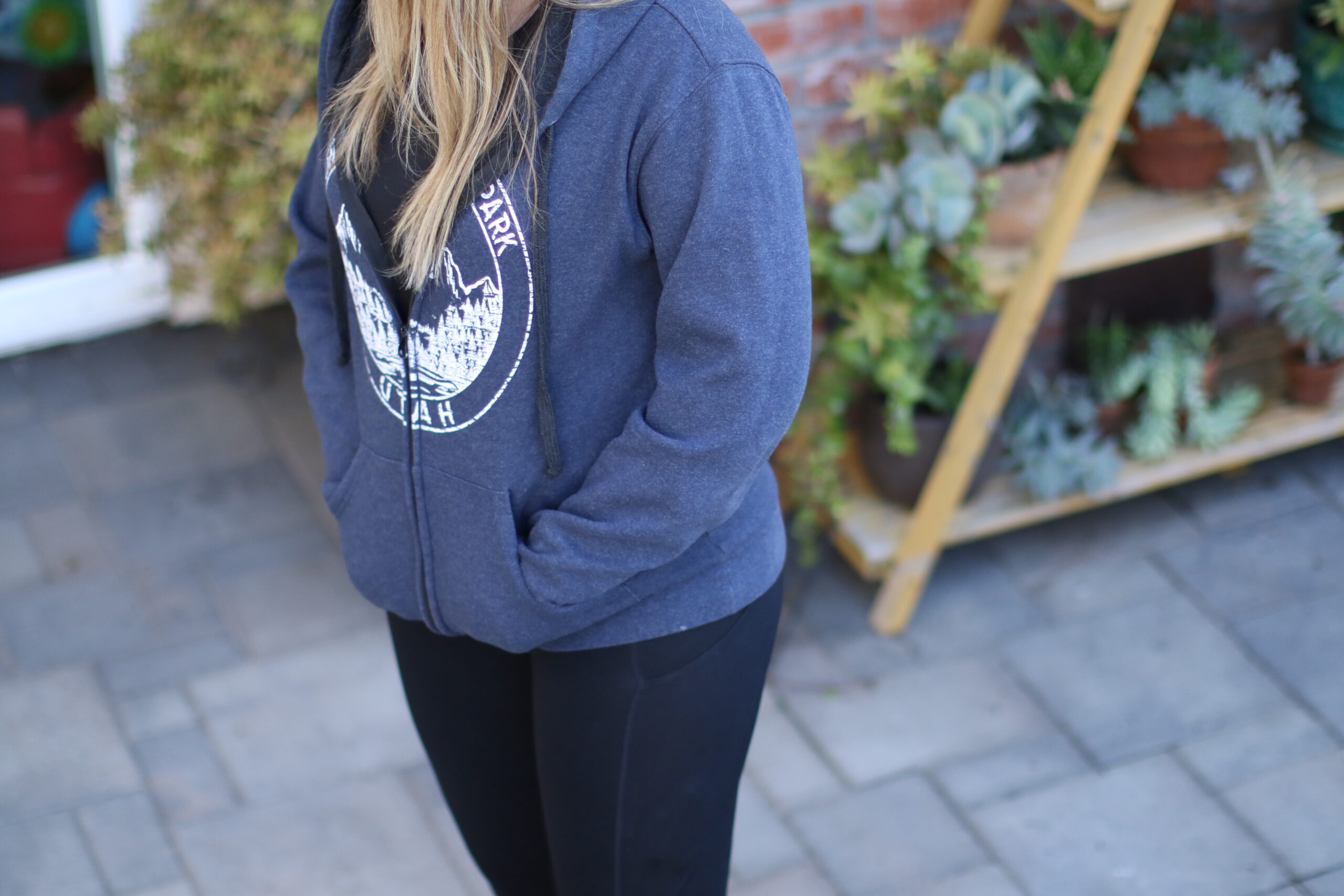 Woman wearing a blue hoodie with her hands in the sweatshirt pockets standing outside on gray pavers in the backyard