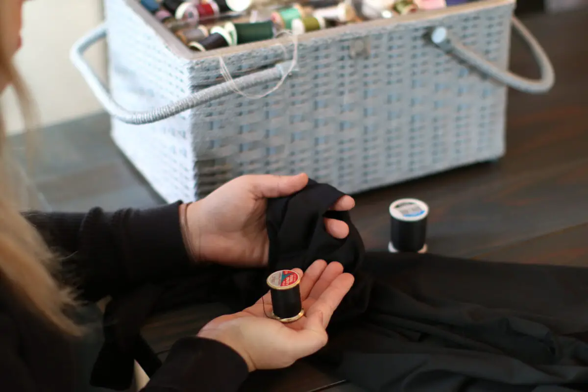 A woman holds black thread fixing a hole in black leggings beside a basket full of sewing tools