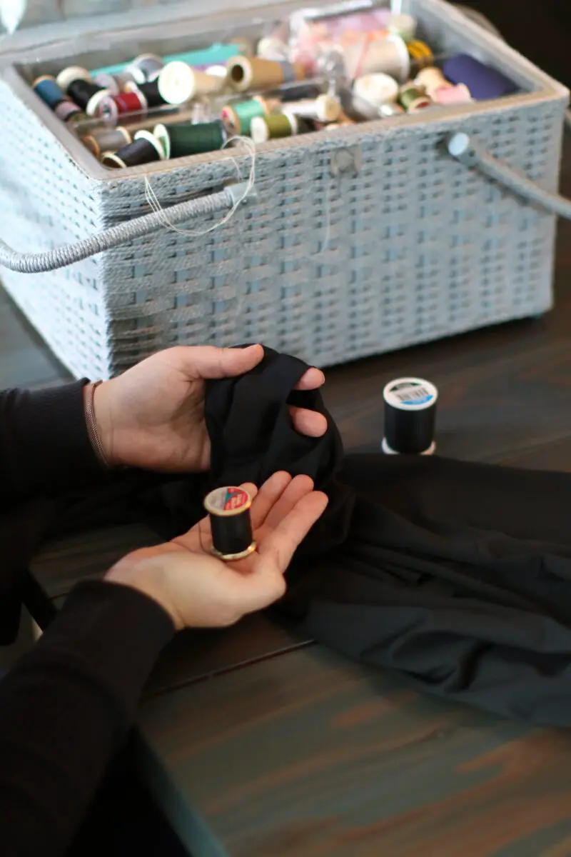 A basket of sewing tools is placed on a wooden table while a person holds a black thread fixing a hole in a black leggings