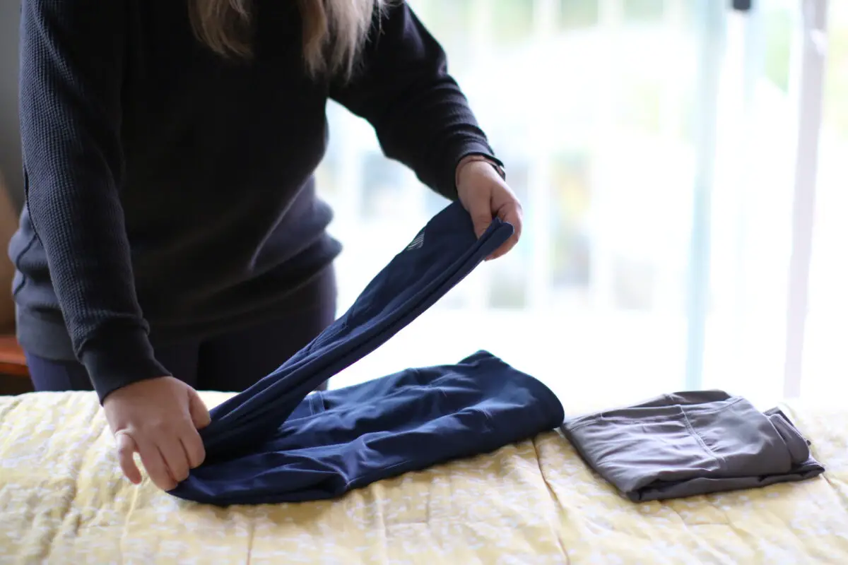 A woman folding blue leggings beside folded gray leggings on the yellow bedsheet of a bed