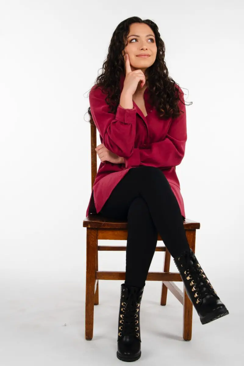 Woman in a red long sleeves and black leggings wearing black boots seated on a brown wooden chair.
