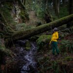 Woman in a yellow jacket and green leggings wearing brown rubber shoes standing in the middle of the forest