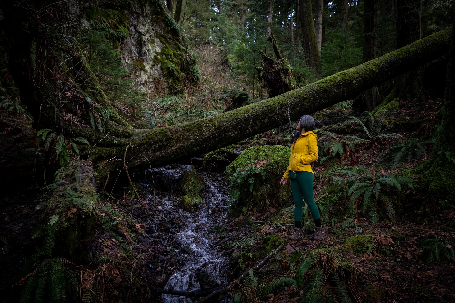 Woman in a yellow jacket and green leggings wearing brown rubber shoes standing in the middle of the forest