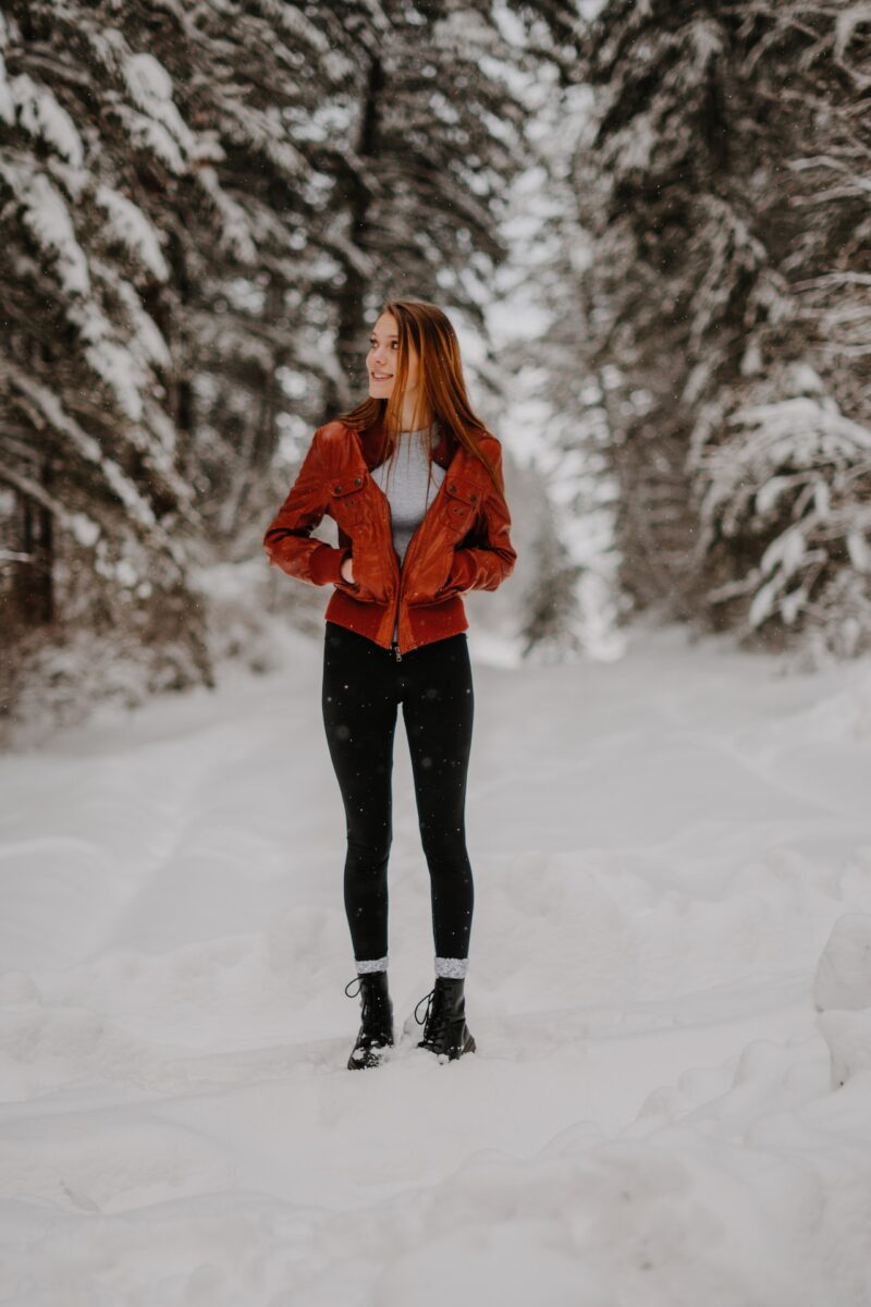 Woman in a gray t-shirt and orange jacket wearing black leggings and black boots standing on a snowy park