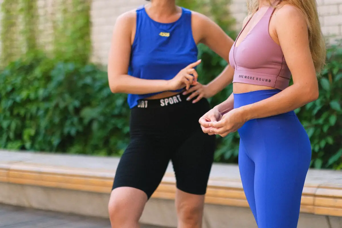 One woman in a pink crop top and blue leggings beside a woman in a blue tank top and black leggings talking in the street