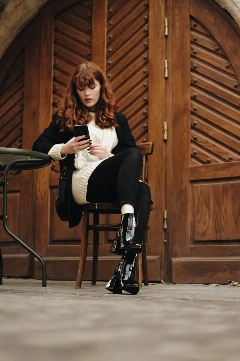 Woman in white knitted long sleeves over a black blazer wearing black leggings and black boots seated on a brown chair using a smartphone