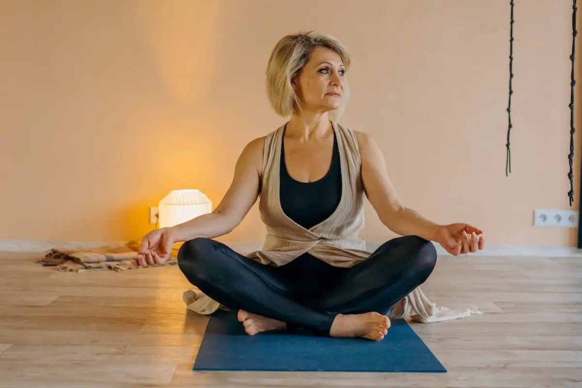 Woman in a black tank top over a brown sleeveless blazer and wearing black leggings while doing a lotus position on a blue yoga mat