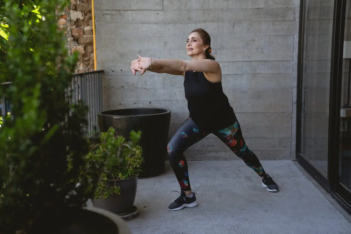 Woman in a black tank top and printed leggings wearing blue rubber shoes doing stretching outside the house