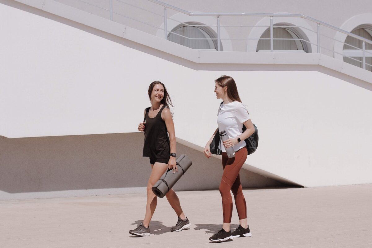 One woman wearing a white t-shirt and brown Capri leggings stands beside the other woman wearing a black tank top and black shorts