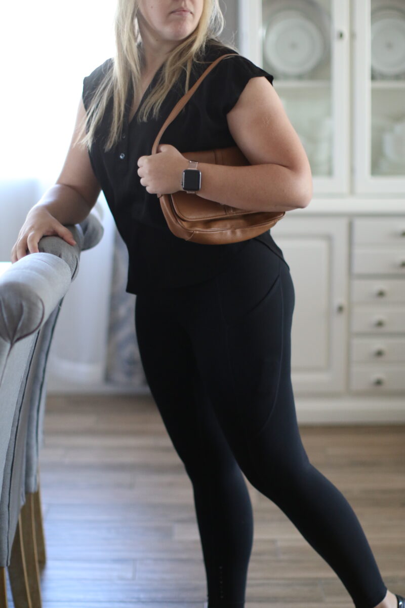 Woman leaning against a chair in the dining room while holding a brown purse and wearing a black blouse and black leggings